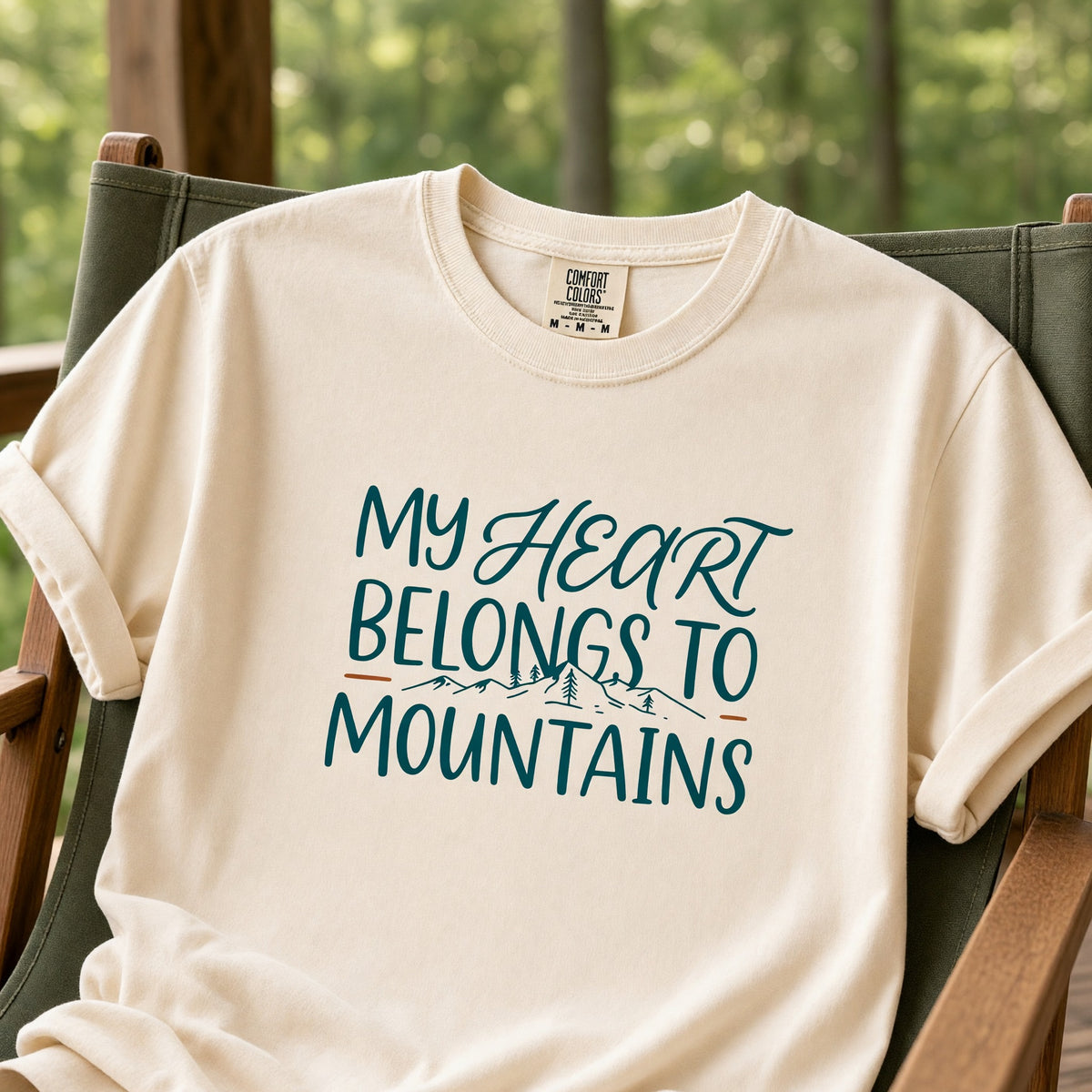 An ivory Comfort Colors t-shirt printed with My Heart Belongs to Mountains, draped over a green canvas camp chair on a porch