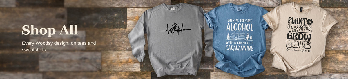 Woodsy graphic tees and a crewneck sweatshirt arranged against rustic barnwood