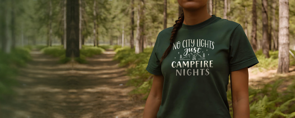 Woman in a forest green t-shirt printed with the Campfire Nights design, standing on a sunlit forest trail