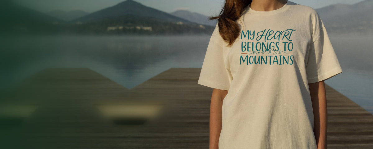 Woman in an ivory t-shirt printed with the My Heart Belongs to Mountains design, standing on a wooden dock at a misty lake