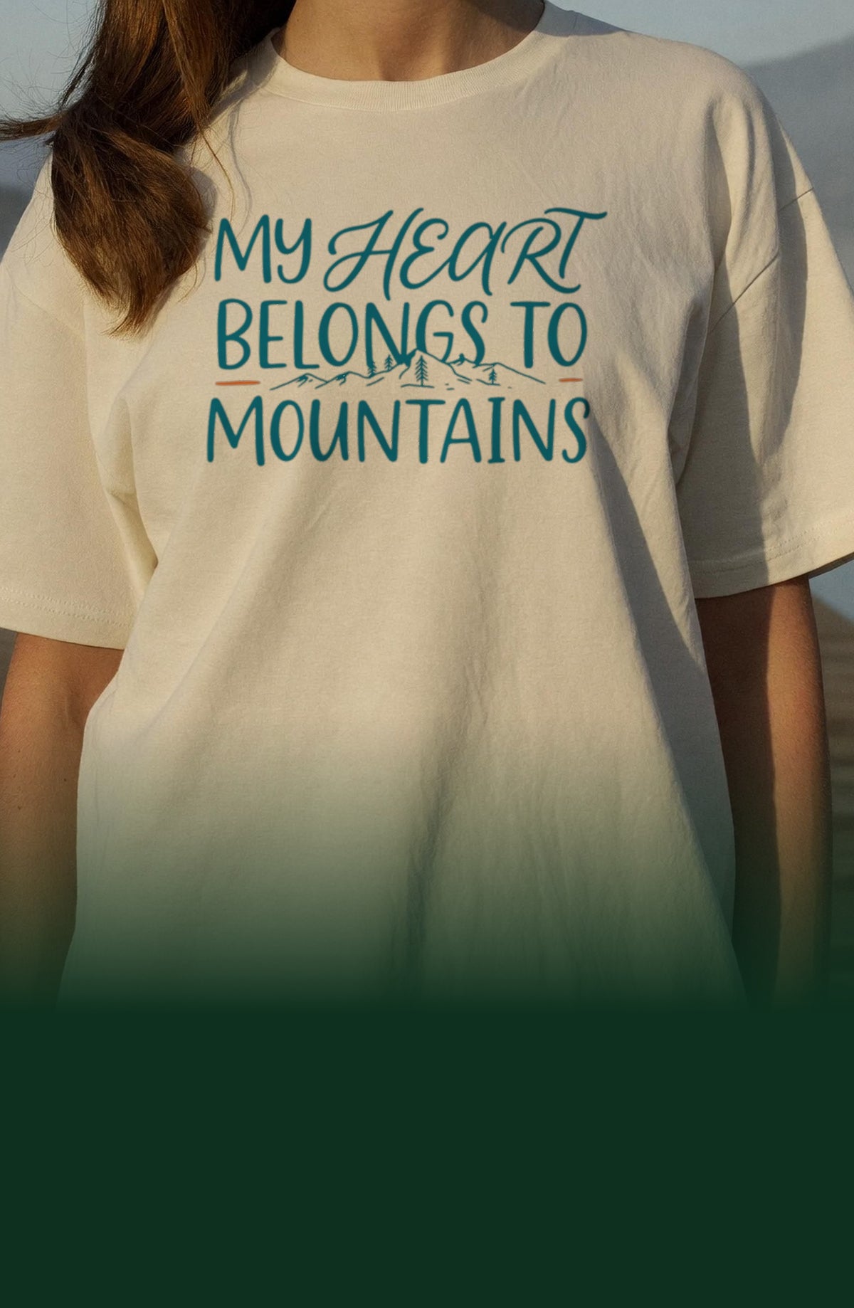 Woman in an ivory t-shirt printed with the My Heart Belongs to Mountains design, standing on a wooden dock at a misty lake